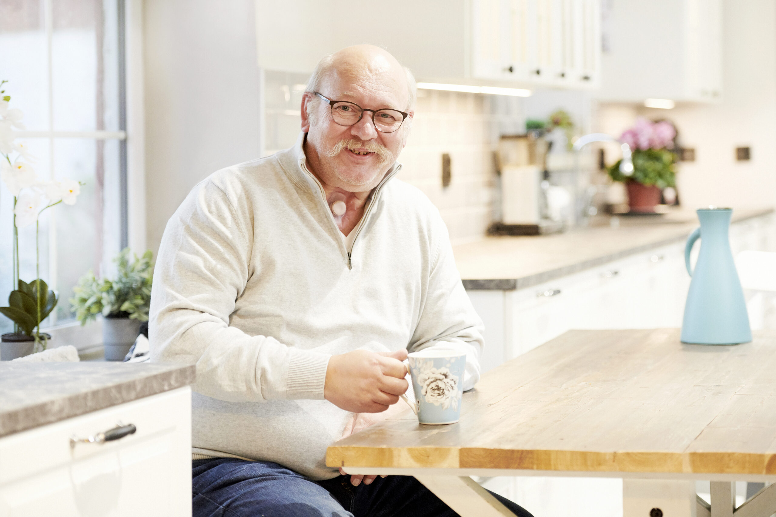 Man with a neck stoma sitting in the kitchen holding a mug using Provox Life HME.