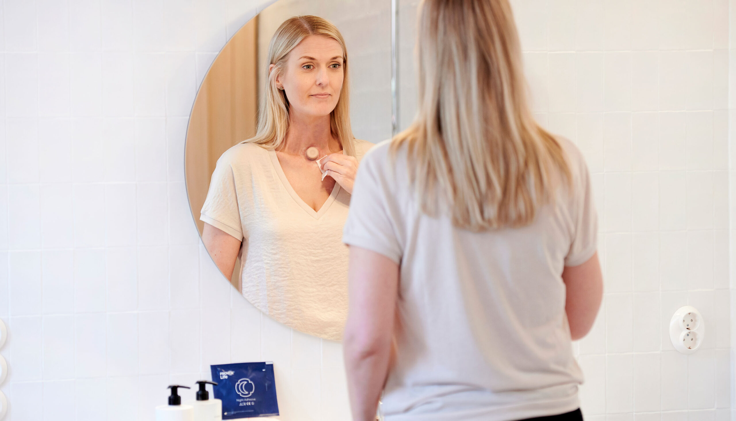 Young woman with a neck stoma looks in the mirror and takes care of her Provox life Home HME.